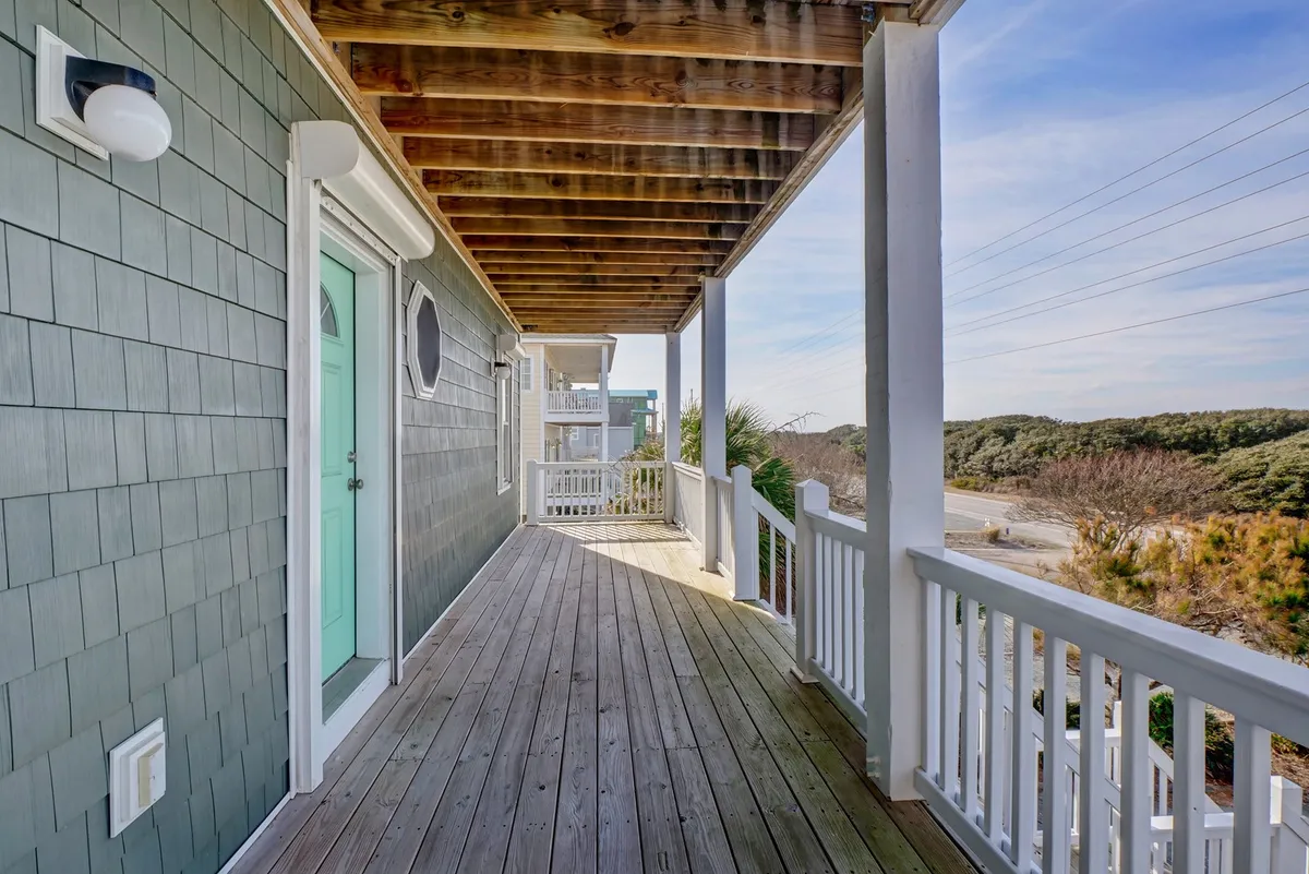 A relaxed exterior deck scene with wooden decking, green siding, white railings, and a door with an oval window. Deck chairs invite guests to unwind and savor the sea breeze, ideal for outdoor meals or drinks at sunset.