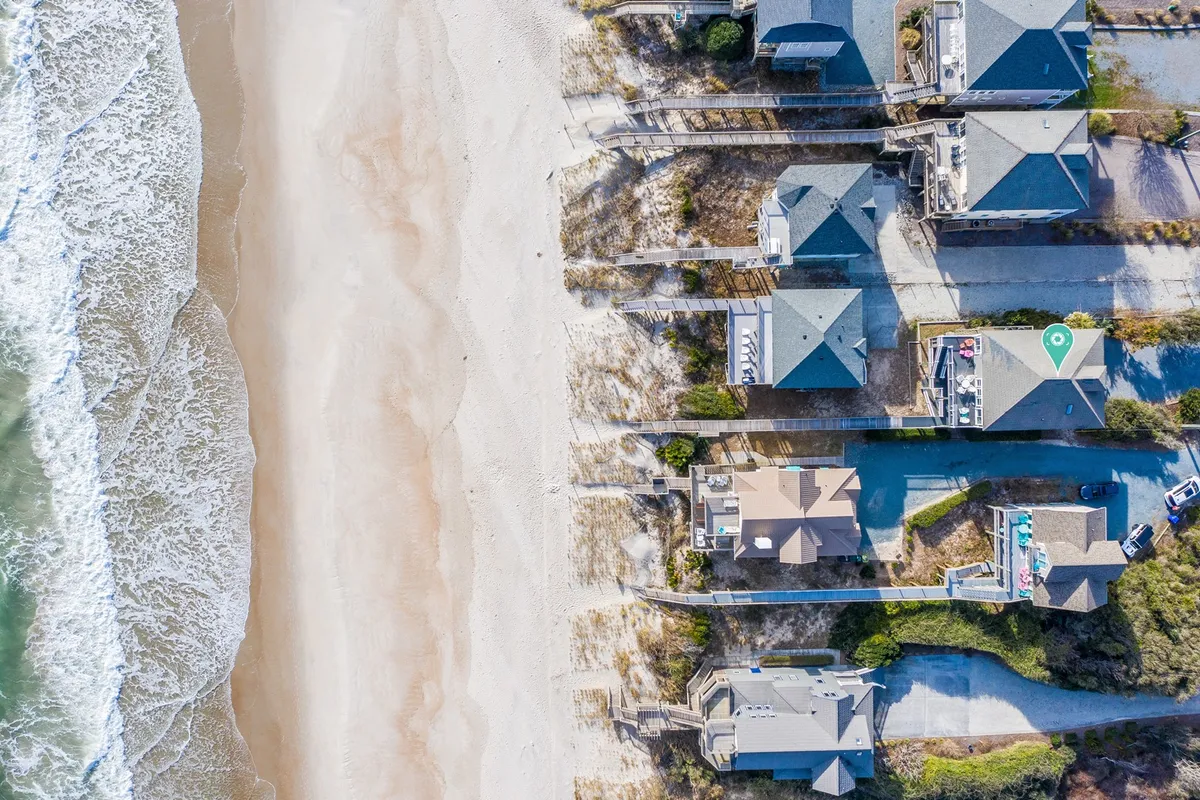 A row of beachfront houses overlooks a sandy beach with gentle waves rolling in. Walking paths lead from the homes down to the shoreline, and the scene is bathed in soft, welcoming light. The space feels open and inviting, ideal for family walks, group gatherings, or water activities.