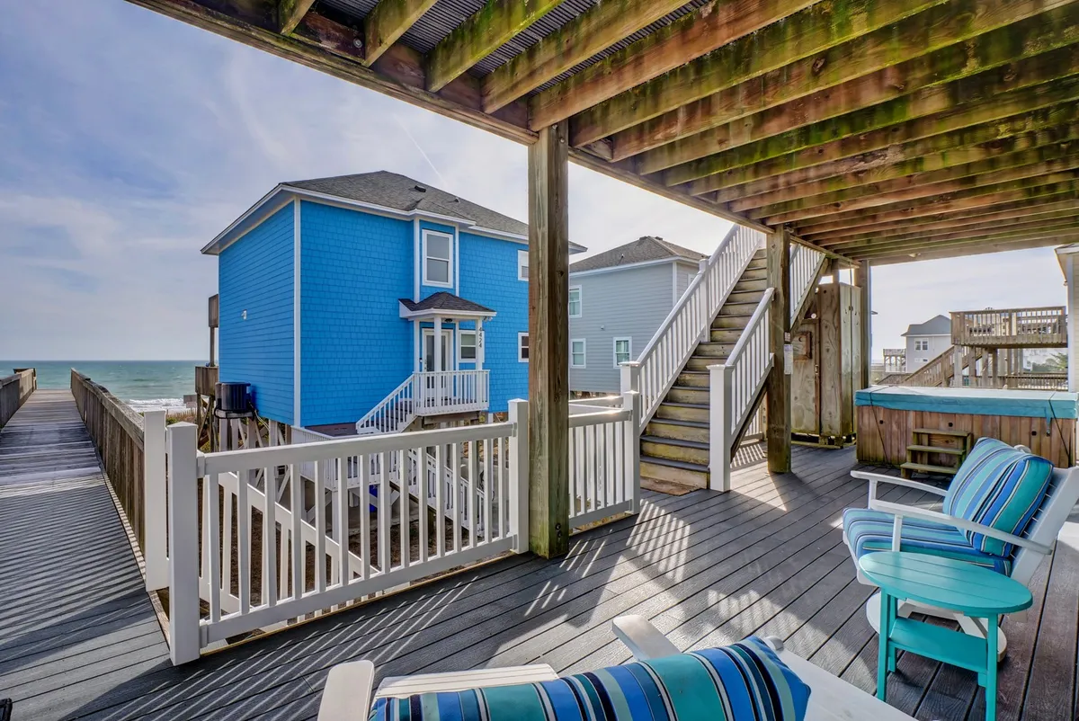 A welcoming deck scene with deck chairs positioned to enjoy the ocean view, the blue exterior of the house, and a staircase leading up to the living spaces. The coastal atmosphere is enhanced by the light and open sky, suggesting both social gatherings and moments of solitude.
