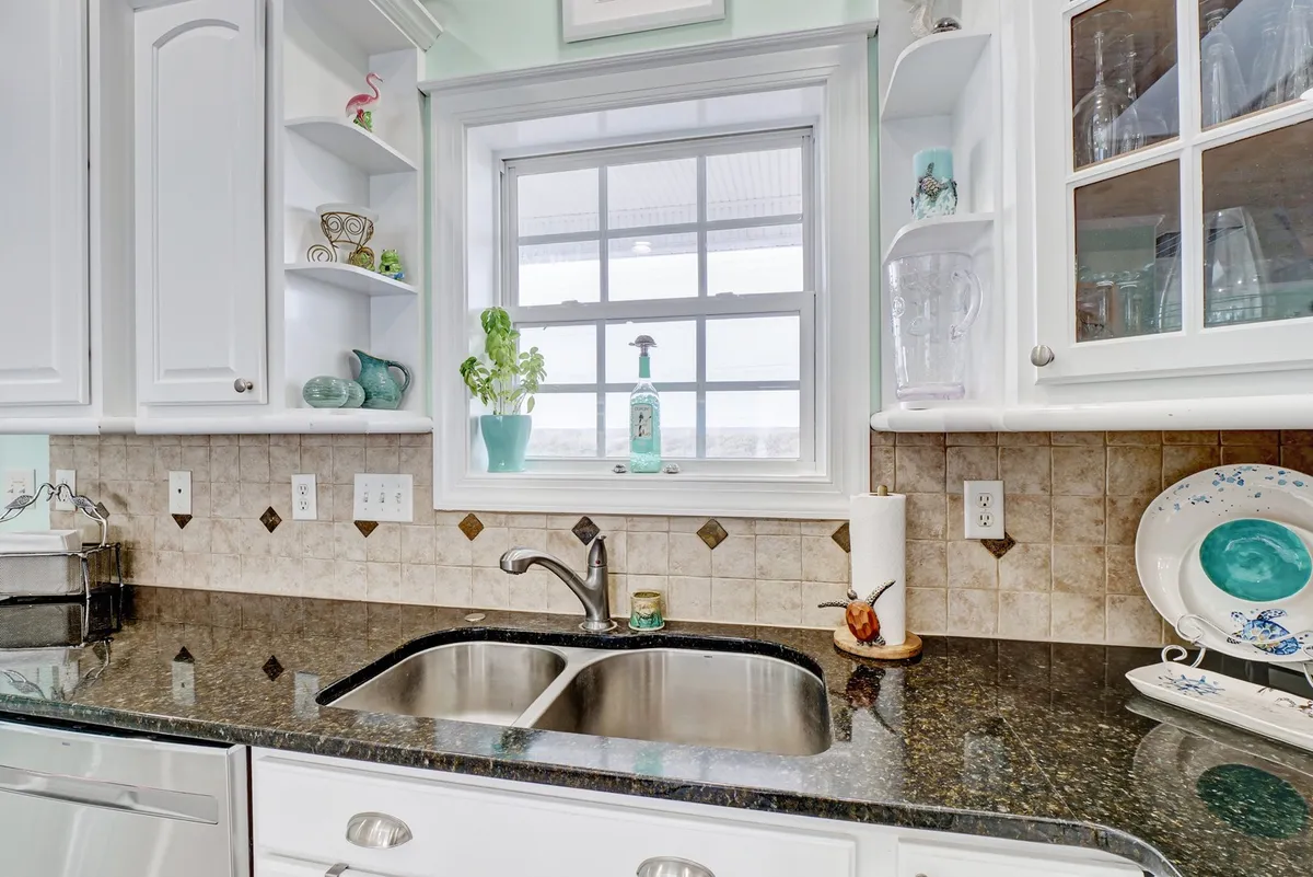 A cheerful kitchen corner with a sunlit window over the sink, potted plants, and decorative items on shelves. The light, inviting palette and open workspace encourage both everyday cooking and special gatherings.