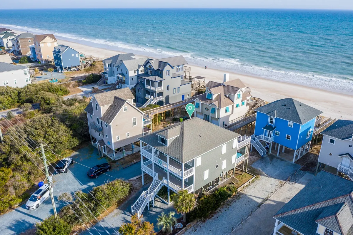 This image showcases a row of beachfront houses along a sandy shoreline, framed by palm trees and walkways. The ocean stretches into the background, inviting visitors to imagine themselves exploring or relaxing by the water. The presence of walkways and nearby shops aligns with the idea of venturing out from the home to nearby local attractions.