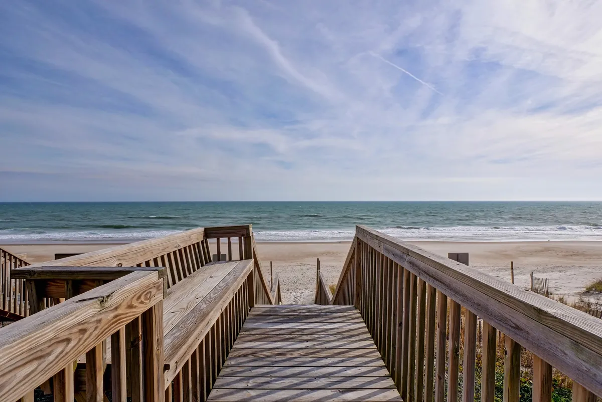 A wooden walkway leads directly over sandy dunes to the ocean, with gentle waves visible in the distance. The view captures both the proximity of the home to the beach and the inviting, open access to the shoreline. The sky is clear, suggesting a peaceful, relaxing day perfect for seaside adventures.