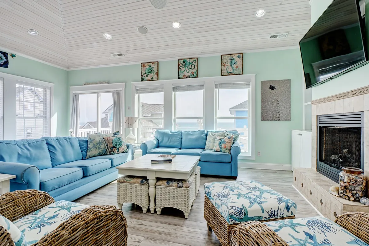 This image showcases a welcoming living area with light blue sofas, woven wicker chairs, and decorative pillows arranged around a coffee table adorned with shells. A television is mounted on the wall, and large windows suggest the presence of natural light and possible water views.