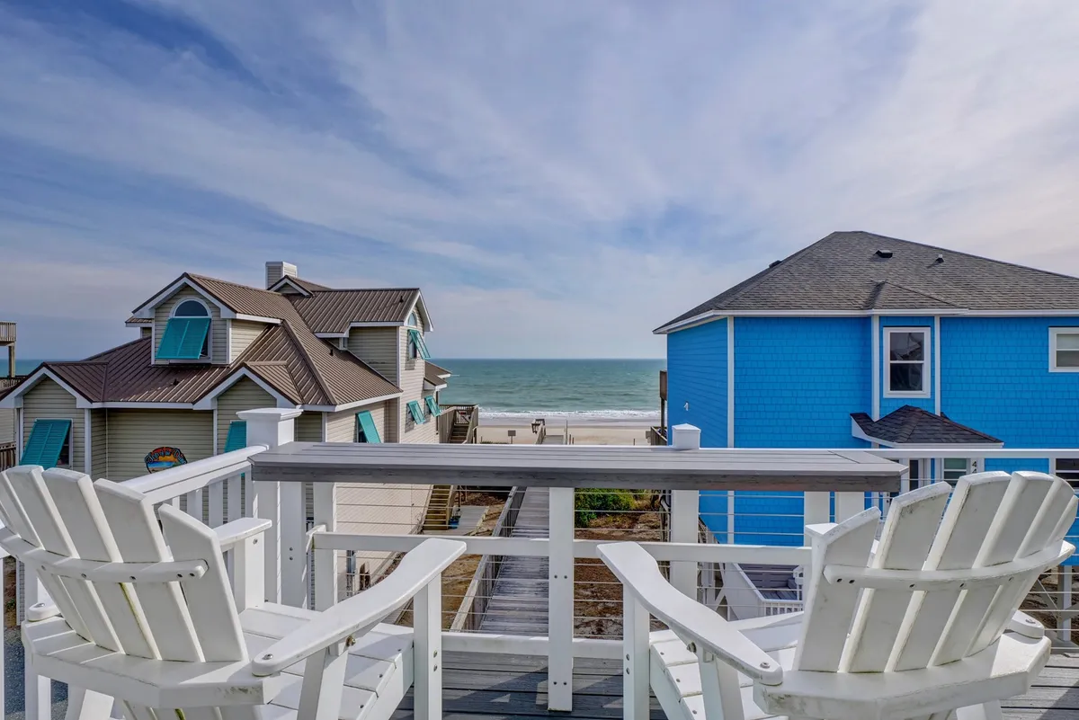 The image showcases a serene deck with inviting Adirondack chairs overlooking the sparkling Atlantic Ocean. Colorful beach houses dot the shoreline in the distance, while the expansive blue sky completes the tranquil seaside scene. The setup encourages relaxation and socializing, with plenty of space to gather and soak in breathtaking ocean views.