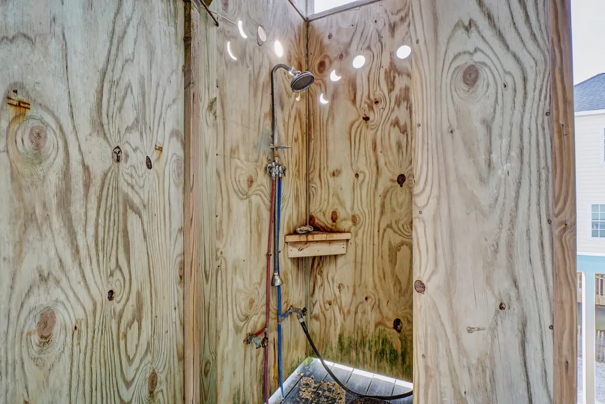 A rustic outdoor shower with wooden walls, a showerhead, and sunlight streaming through small openings. The natural light and open-air feel offer a refreshing post-beach rinse while maintaining privacy.
