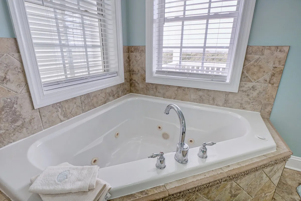 A tranquil bathroom interior featuring a jacuzzi-style tub bathed in soft natural light from nearby windows, surrounded by textured wall tiles. The setting exudes a calming, spa-like ambiance perfect for relaxing after a day outdoors.
