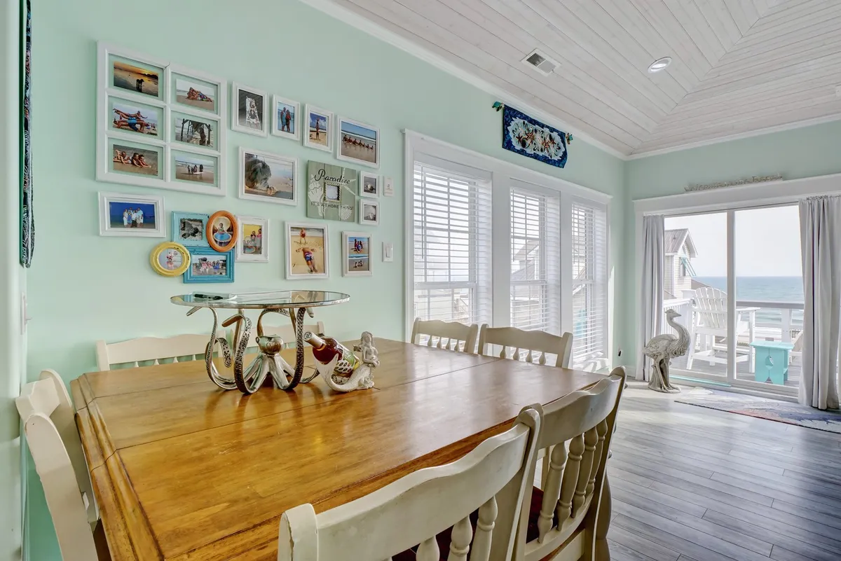 A cozy dining area with a sturdy dining table, a gallery wall with family photos, and expansive windows offering views of the ocean. The space is filled with sunlight, inviting guests to enjoy meals together while taking in the scenic surroundings.