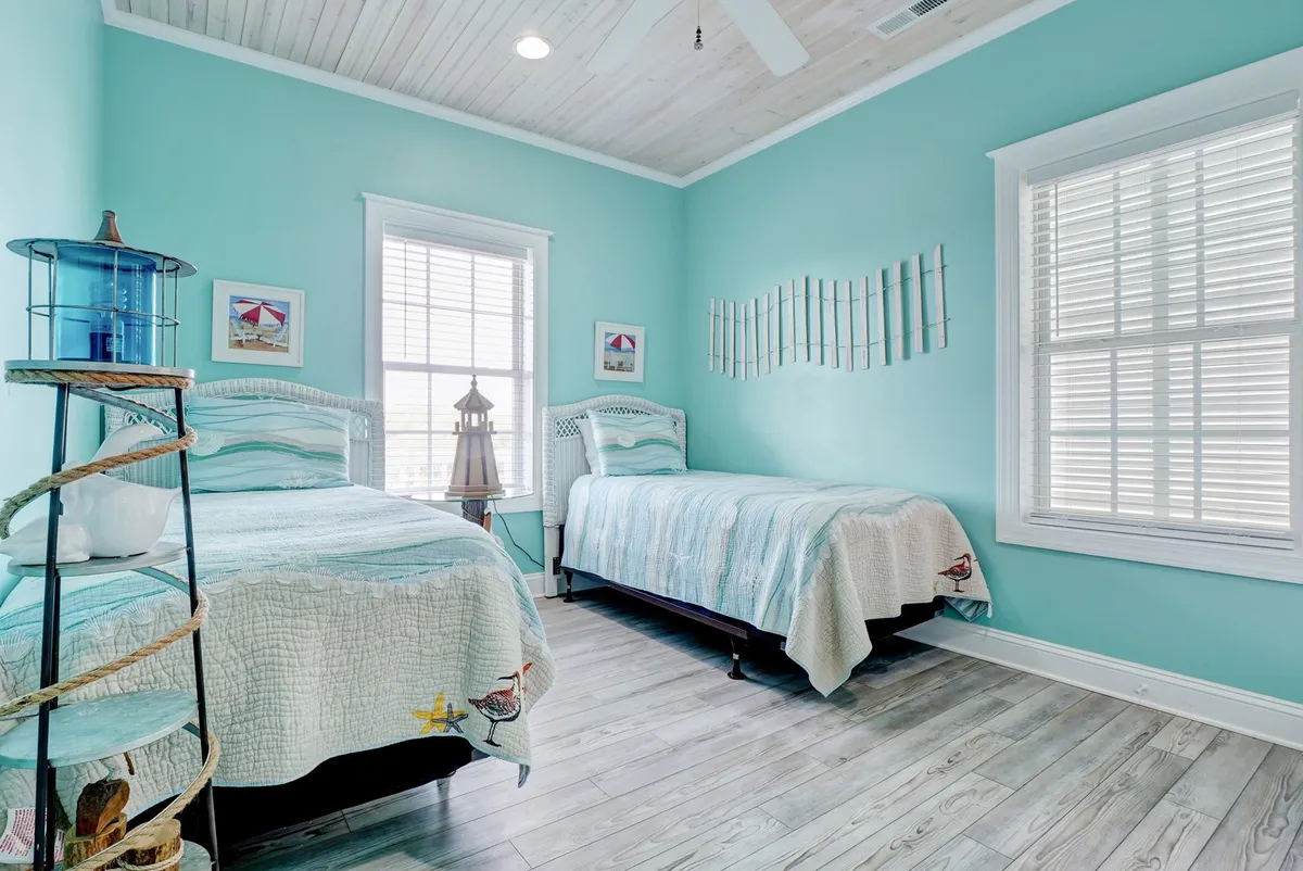 A cheerful bedroom featuring two twin beds with turquoise walls, a decorative lantern, and nautical-themed accents. Sunlight streams through the window, creating a calm ambiance ideal for solo or shared relaxation.