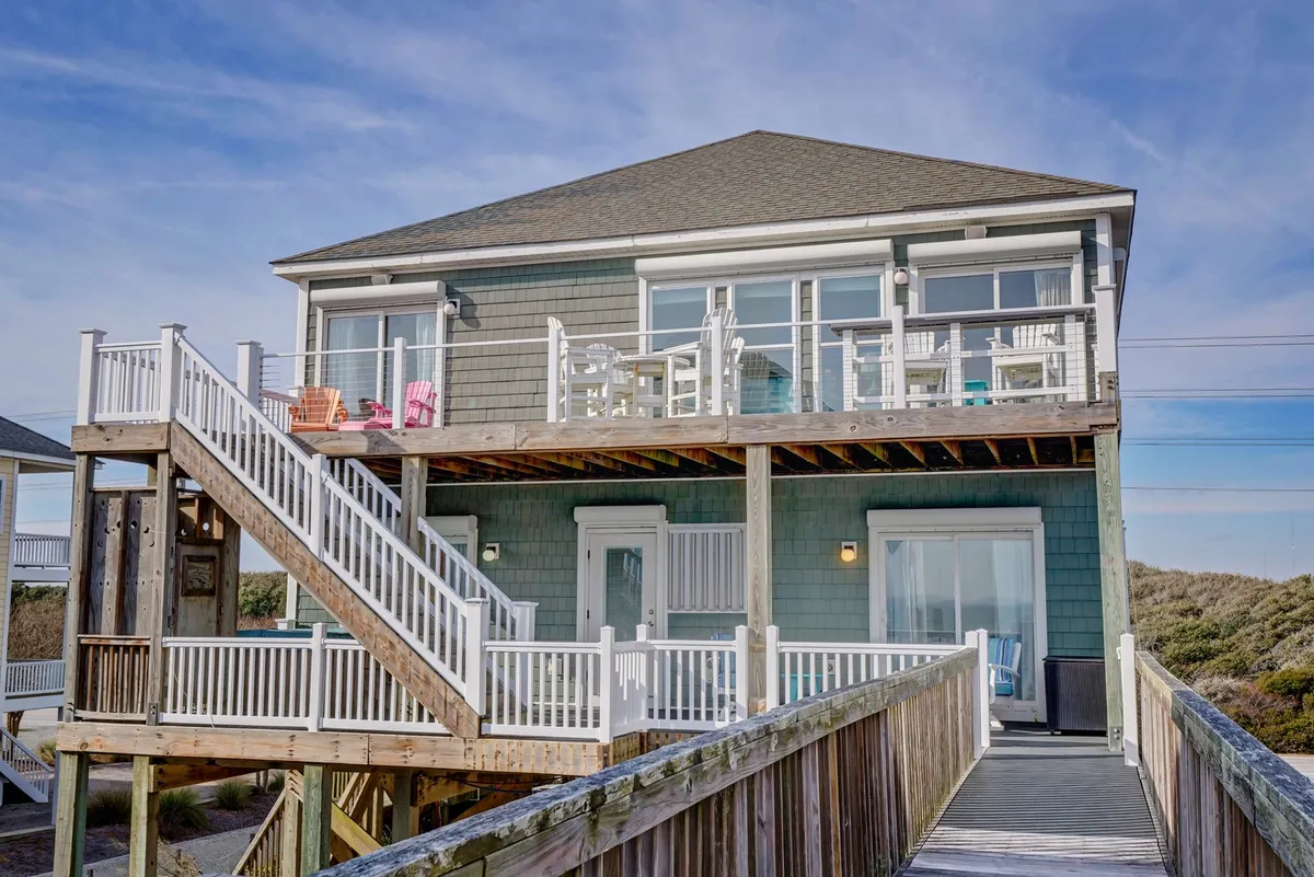 A two-level deck extends from the house, complete with outdoor seating and large windows. The staircase leads directly to the entrance, and there’s ample space for relaxing, entertaining guests, or playing games outside. The mood is welcoming and relaxed, set against a backdrop of open skies.