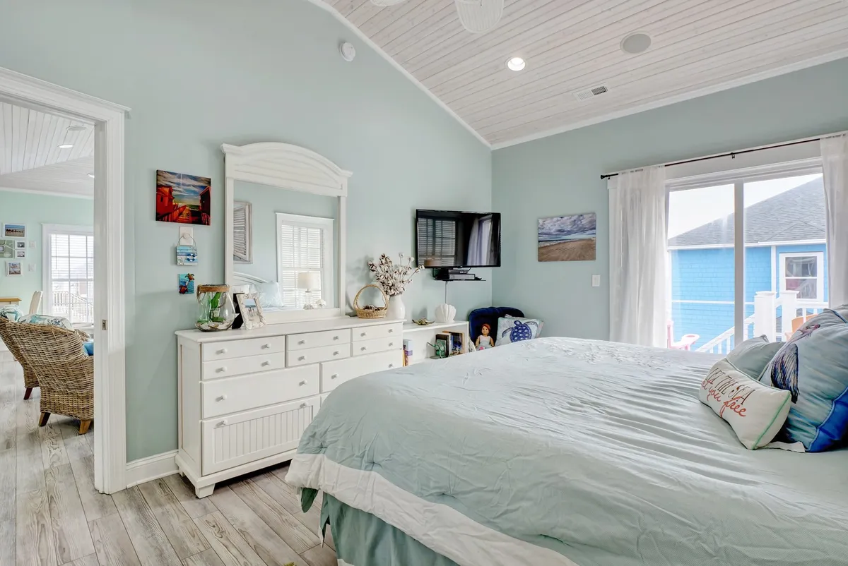 A tranquil bedroom with a king-sized bed adorned in light blue bedding, a white dresser with mirror, and large windows that bathe the space in natural light. Decorative pillows with inviting text add a thoughtful touch, creating an environment that feels both restful and welcoming.