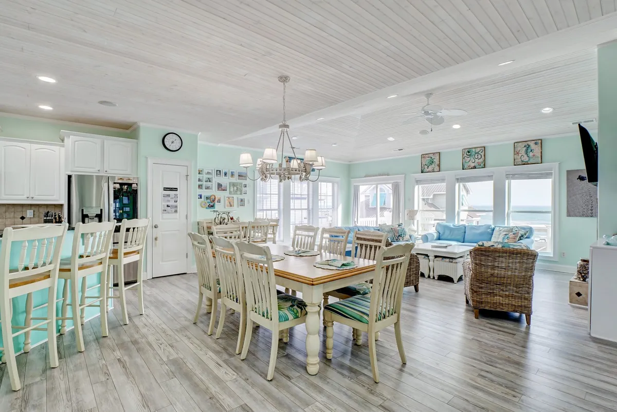 A bright, open-plan space featuring a dining table next to a kitchen island, with large windows and a comfortable seating area. The room is filled with natural light and offers a cheerful atmosphere ideal for group meals and conversations.
