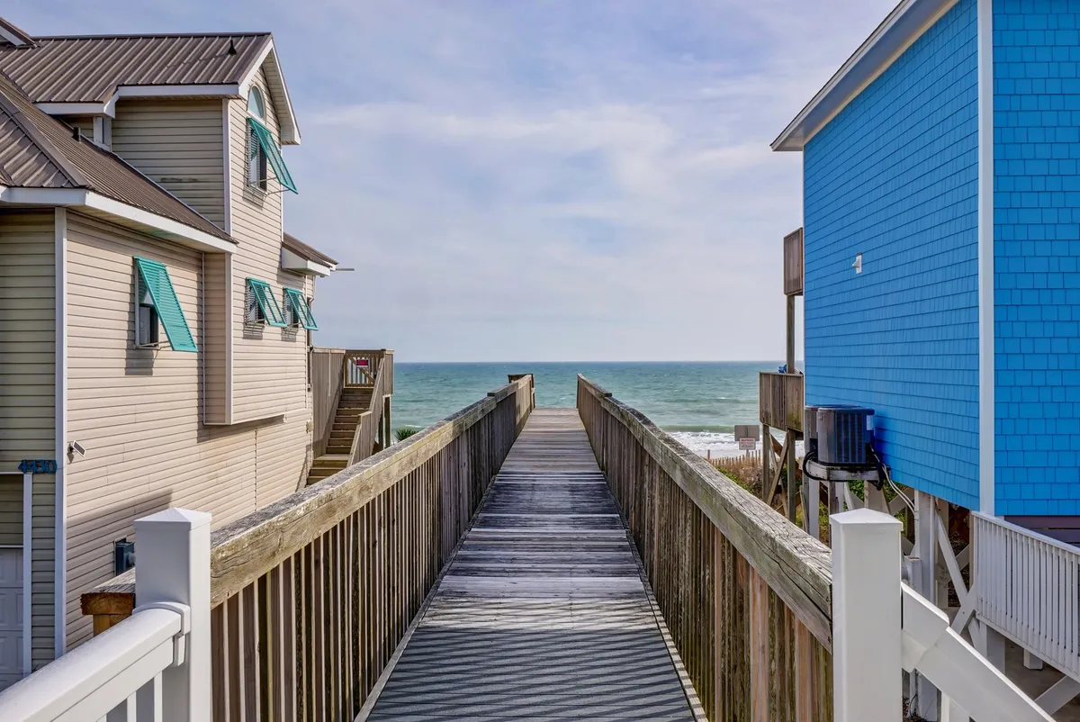 A relaxing wooden walkway extends from a blue house directly to a wide, sandy beach with unobstructed ocean views. The composition connects the home seamlessly to the shoreline, echoing the concept of beach adventures just outside the door. The tranquil setting suggests both daytime fun and evening relaxation.